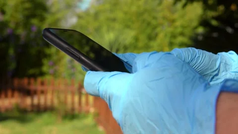 Closeup of hands wearing blue gloves holding a smartphone in a sunny garden. Stock Footage 299082808