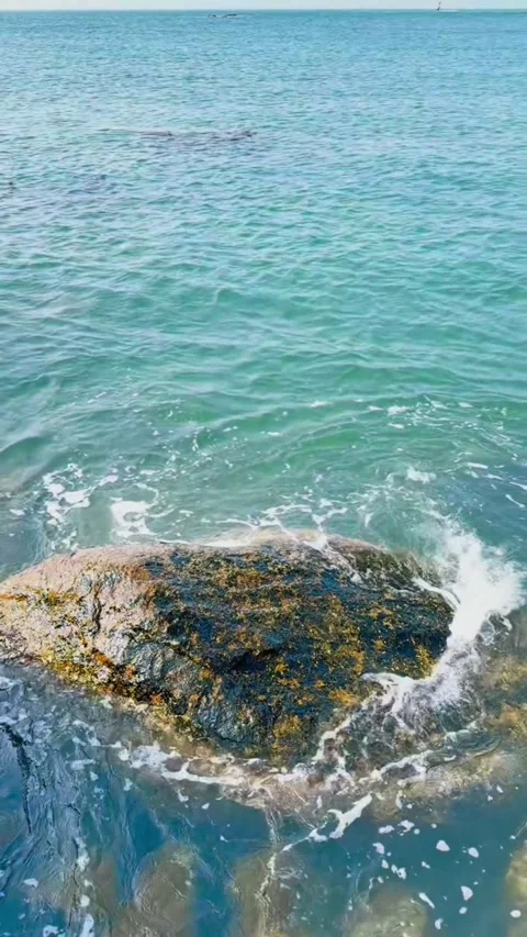 Closeup of a large rock partially submerged in the clear blue ocean water with Vídeos de archivo 334353568