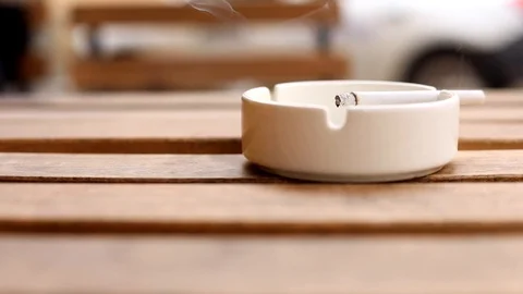 Closeup macro view of cigarette in white ashtray on wooden desk outdoors on blur 스톡 동영상 118506435