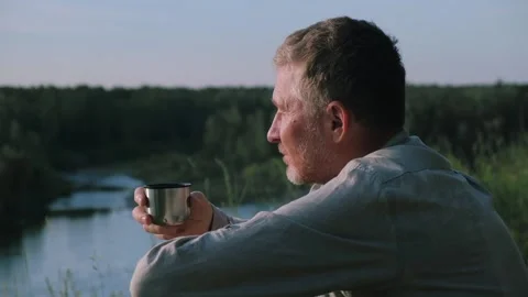 Closeup man drink tea from mug on riverbank and looks thoughtfully into distance Stock-Footage 231990909