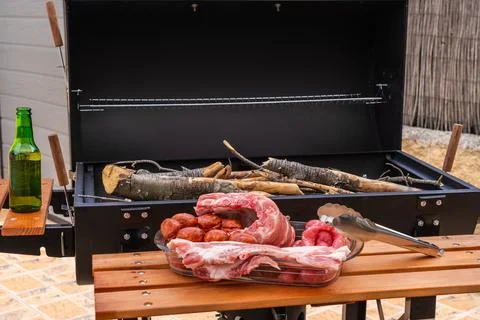 Closeup shot of an array of raw meats near an outdoor grill with wood 스톡 사진