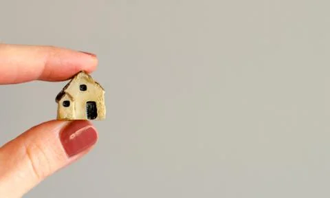 Closeup of tiny plastic house holding in the woman's fingers on blurred grey  Stock Photos