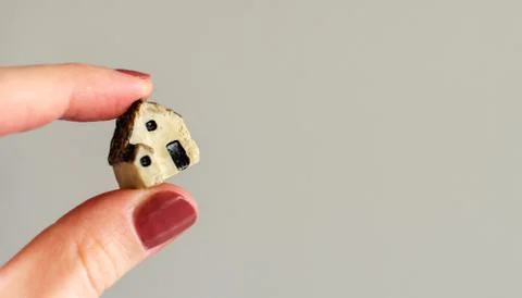 Closeup of tiny plastic house holding in the woman's fingers on blurred grey  Stock Photos