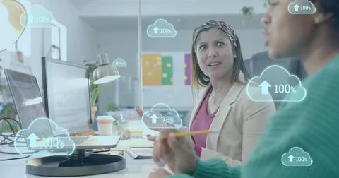 Cloud icons uploading data over diverse female colleagues discussing at computer Stock Photos
