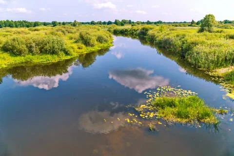 The clouds are reflected in the blue river, dividing the green grassy terrain Stock Photos