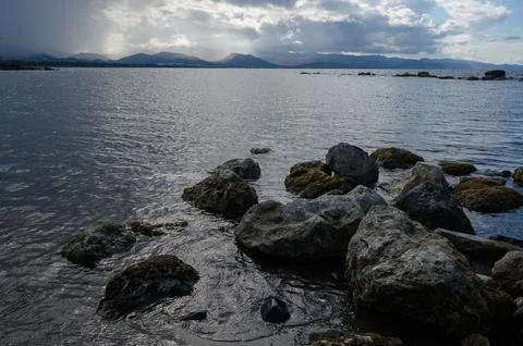 Clouds gather over tranquil waters, as rocks line the shore in a serene coa.. Stock Photos