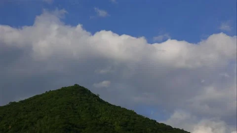 Clouds move steadily across bright blue sky above a lush green mountain covered Stock Footage 322166593