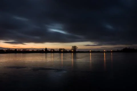 Cloudy dark clouds after sunset on a frozen lake and reflecting lights on the Stock Photos