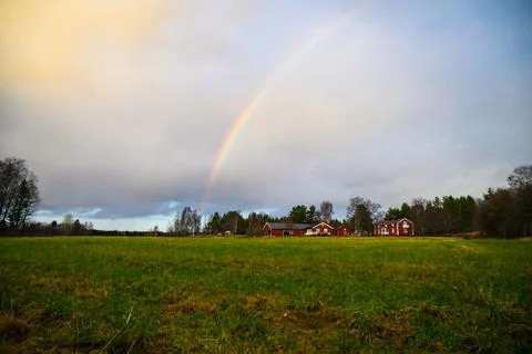 A cloudy landscape view of open field with cottages and rainbow in the sky in Stock Photos