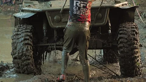 A co-pilot stands in thick mud next to a dirty SUV, reeling in a winch cable Stock Footage 345497190
