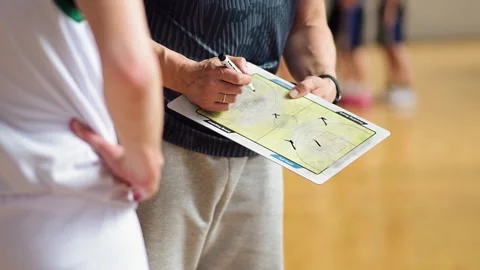 Coach makes notes during practice at a sports gym with players in the background Stock Footage 340357009