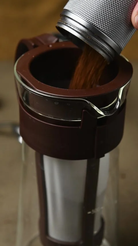 Coarse coffee grounds are poured from a metal grinder into a cold brew filter in Stock Footage 346743728