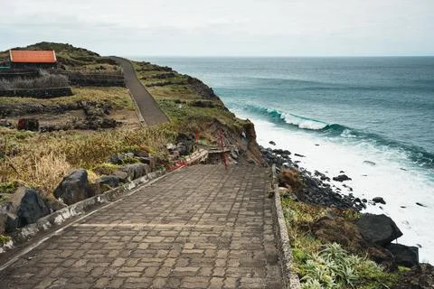 Coastal Path Leading to Ocean with Waves, Rocky Shore, and Distant Building.. Foto stock
