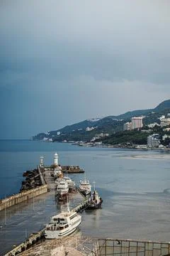 Coastal pier with multiple boats docked, lighthouse at end, mountainous bac.. Stock Photos