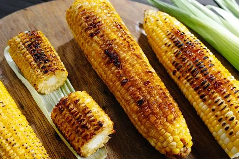 A cob of corn roasted over charcoal on a wooden board. Whole corn cobs 库存照片