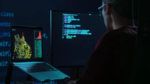 Coder works at desk in a dark office with programming code lines running behind Видео 345446316