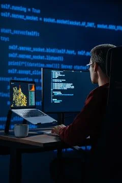 Coder works at desk in a dark office with programming code lines running behind Stock Photos