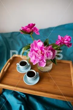 Coffee and pink peonies on a wooden tray in bed with bright linens. Stock Photos