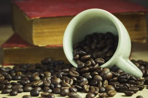 Coffee bean in tilted ceramic cup and old book laid behind on wooden table. Stock Photos