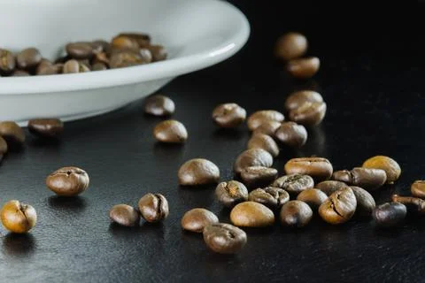 Coffee beans on a black surface, next to a white saucer on which are coffee b Stock Photos