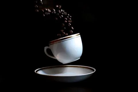 Coffee beans fall into a coffee cup hovering over a saucer on a dark backgrou Stock Photos