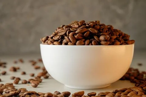 Coffee beans piled high in a white bowl with scattered beans around Foto stock