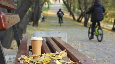 Coffee break on empty bench in autumn city park and two blurred boys on bicycles 스톡 동영상 140659731