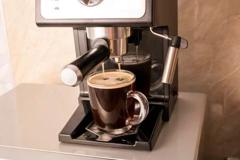Coffee is poured from the coffee machine into a transparent glass glass Stock Photos