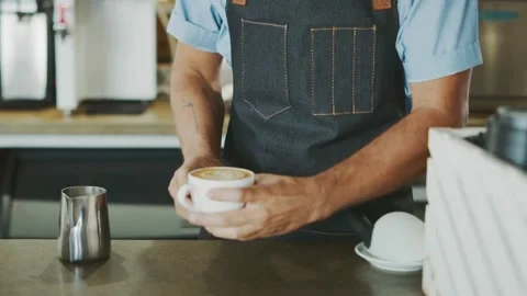 Coffee shop worker in apron pours milk creating cafe latte art in cup. Stock-Footage 334648645