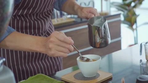 Coffee shop worker in apron pours milk creating cafe latte art in cup. 動画素材 334649853
