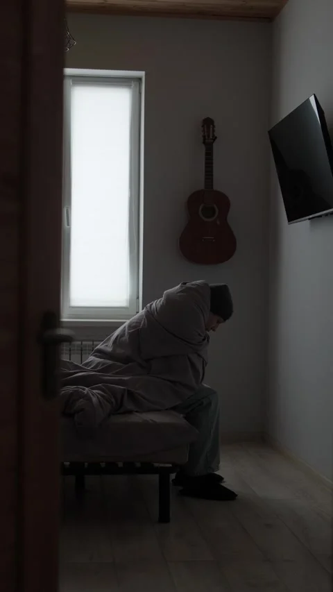 Cold apartment documentary, man sitting on bed under blanket seen through Stock Footage 329626610
