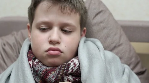 A cold boy coughs. He has the flu, he's sitting on the couch in a scarf 스톡 동영상 97274502
