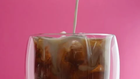 Cold brew with ice and creamy swirl slowly mixes in rotating glass against pink Stock Footage 305863427