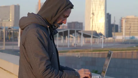 Cold hooded male hacker quickly typing code on laptop overlooking downtown Stock Footage 166613637