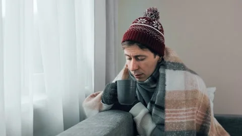 Cold man wrapped in a blanket and winter hat drinks hot tea at home. Concept of  Stock Footage 341880817