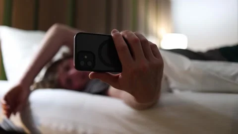 College student lying in bed holding smartphone close to face scrolling social m Stock Footage 318778768