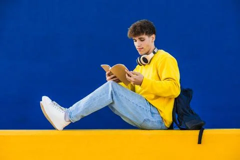 College student reading a book while sitting on a yellow wall with blue Stock Photos
