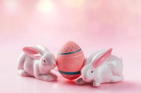 Colored decorated egg in small basket and porcelain rabbits on pink background. Stock Photos