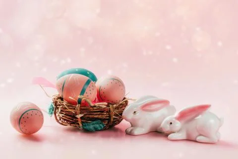 Colored decorated eggs in small basket and porcelain bunnies on pink background. Stock Photos