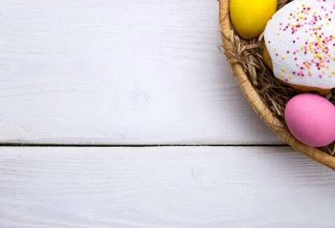 Colored easter eggs and little easter cake in nest beasket on white wooden ba Stock Photos