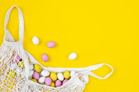 Colored Easter eggs in a mesh bag on a yellow background, flat lay. Stock Photos