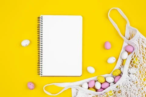 Colored Easter eggs in a mesh bag and a blank notepad on a yellow background. Stock Photos
