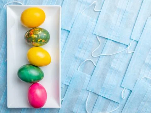 Colored Easter eggs on a white plate on a background of blue medical masks. Stock Photos