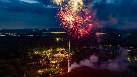 Colorful Fireworks Exploding Over A Well-Lit Estate At Night, Surrounded By.. Stock Photos