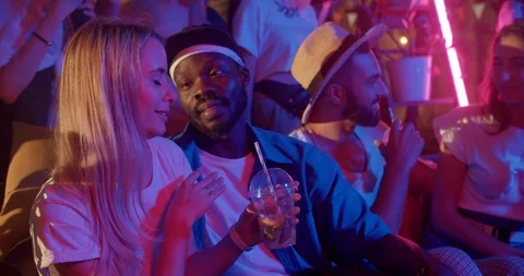 At colorful light multi-ethnic young friends chatting enjoying cocktails at Stock Footage 115392389