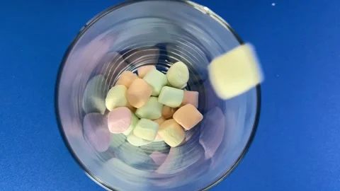 Colorful marshmallow pieces falling into clear glass on blue background Stock Footage 343968459