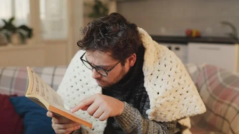 Comfortable cold day Guy with eyeglasses engrossed in a book, wrapped in a duvet Stock Footage 281594924