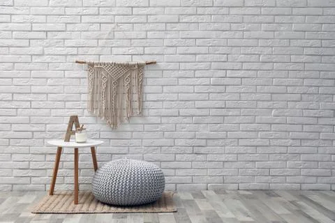 Comfortable knitted pouf, table and decor elements near white brick wall indo Stock Photos