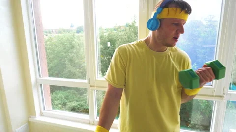 Comic funny athlete with dumbbells pumps a muscle while listening to music Stock Footage 281376572
