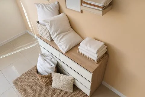 Commode with stack of clean bed sheets, baskets and pillows near beige wall i Stock Photos